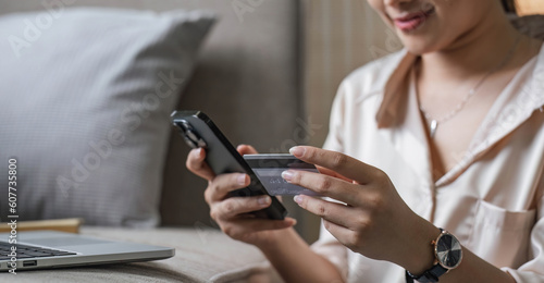 Young smiling Asian woman using smartphone buying online shopping by credit card while lying on sofa when relax in living room at home. Lifestyle latin and hispanic ethnicity women at house concept.