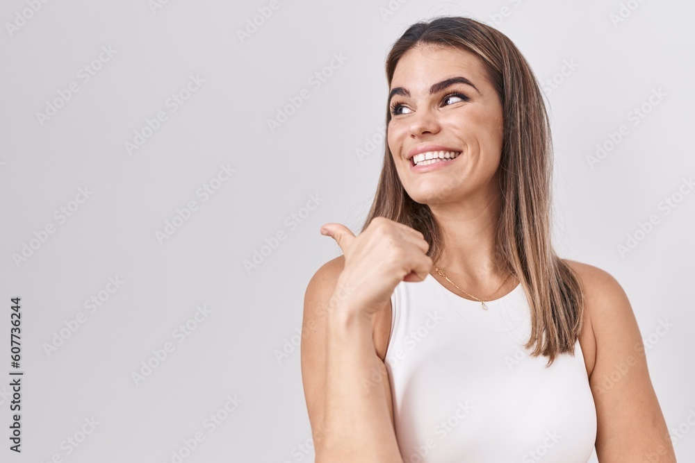 Hispanic young woman standing over white background smiling with happy face looking and pointing to the side with thumb up. Hispanic young woman standing over white background smiling with happy face looking and pointing to the side with thumb up.