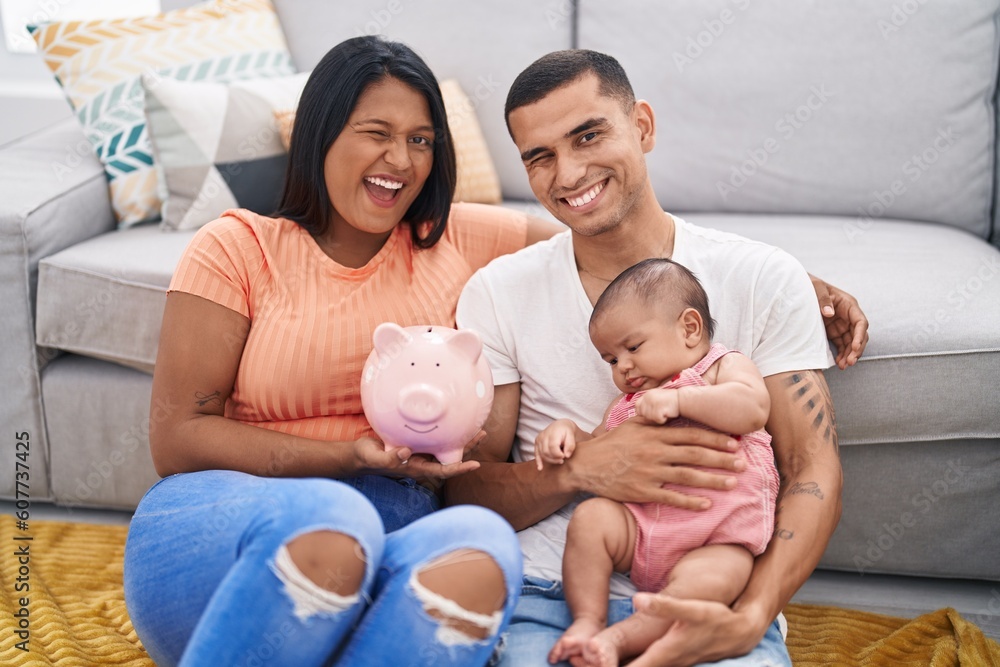 Young hispanic couple with baby holding piggy bank winking looking at the camera with sexy expression, cheerful and happy face. Young hispanic couple with baby holding piggy bank winking looking at the camera with sexy expression, cheerful and happy face.