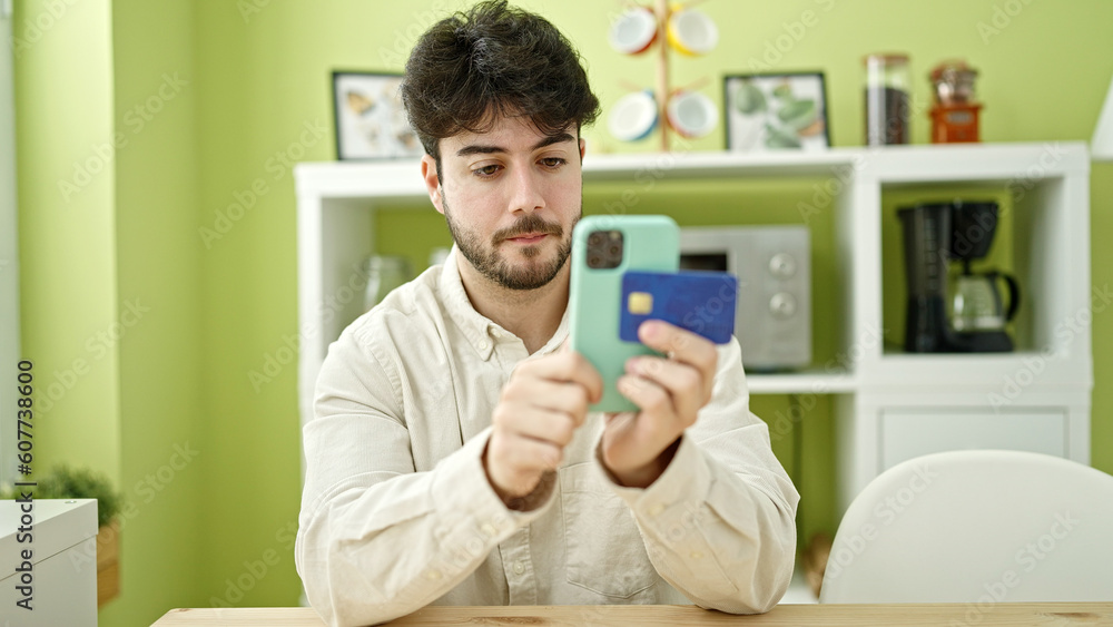 Young hispanic man shopping with smartphone and credit card sitting on table at dinning room Young hispanic man shopping with smartphone and credit card sitting on table at dinning room