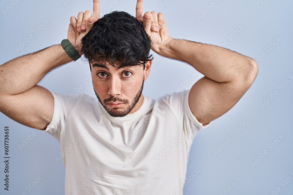 Hispanic man with beard standing over white background doing funny gesture with finger over head as bull horns Hispanic man with beard standing over white background doing funny gesture with finger over head as bull horns