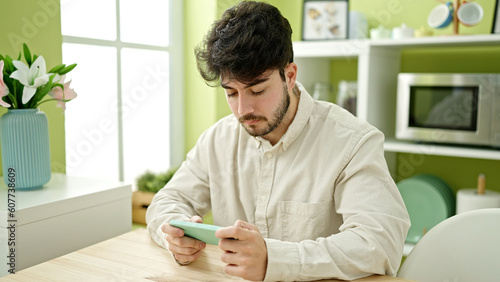 Young hispanic man watching video on smartphone sitting on table at dinning room