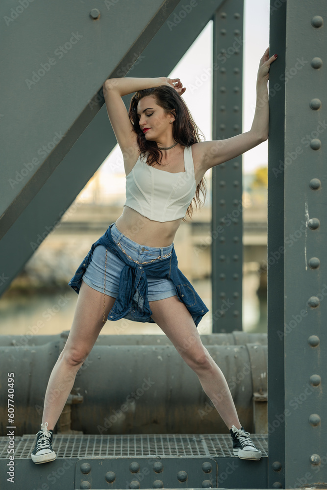 Beautiful girl with red lipstick and dressed in a white top and short jeans posing with her arms resting on the beams of an industrial bridge. Beautiful girl with red lipstick and dressed in a white top and short jeans posing with her arms resting on the beams of an industrial bridge.