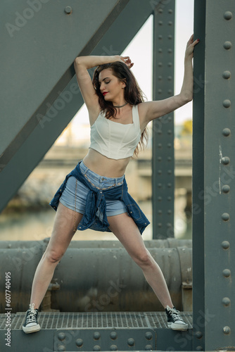 Beautiful girl with red lipstick and dressed in a white top and short jeans posing with her arms resting on the beams of an industrial bridge.
