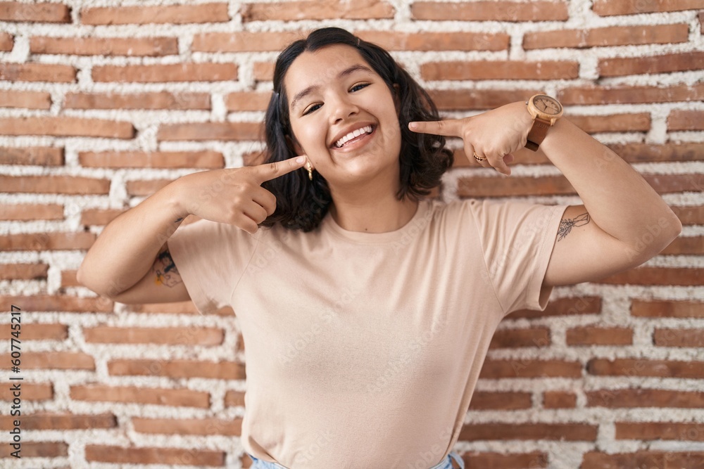 Young hispanic woman standing over bricks wall smiling cheerful showing and pointing with fingers teeth and mouth. dental health concept. Young hispanic woman standing over bricks wall smiling cheerful showing and pointing with fingers teeth and mouth. dental health concept.