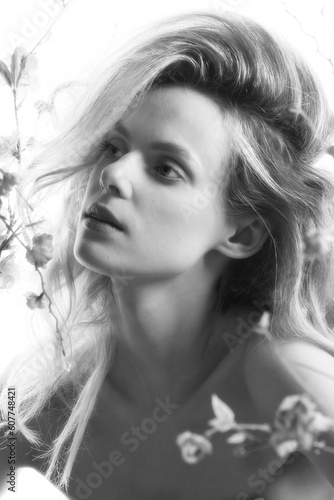 Beauty and make-up concept. Beautiful woman with big wavy hair and make-up studio portrait. Model surrounded with flower twigs with blossoms and naked shoulder looking away. Black and white image