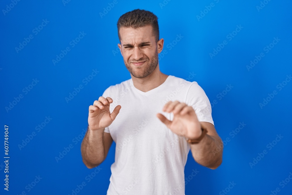 Naklejka premium Young caucasian man standing over blue background disgusted expression, displeased and fearful doing disgust face because aversion reaction.
