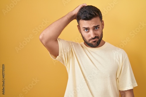 Handsome hispanic man standing over yellow background confuse and wondering about question. uncertain with doubt, thinking with hand on head. pensive concept. © Krakenimages.com