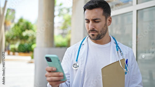 Young hispanic man doctor holding medical report using smartphone at hospital