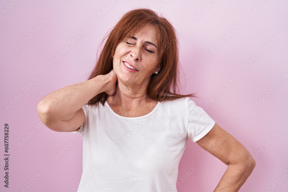 Middle age woman standing over pink background suffering of neck ache injury, touching neck with hand, muscular pain Middle age woman standing over pink background suffering of neck ache injury, touching neck with hand, muscular pain