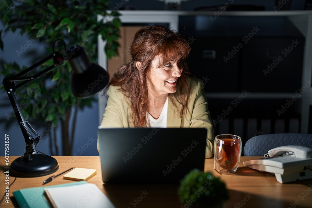 Middle age hispanic woman working using computer laptop at night looking away to side with smile on face, natural expression. laughing confident. Middle age hispanic woman working using computer laptop at night looking away to side with smile on face, natural expression. laughing confident.