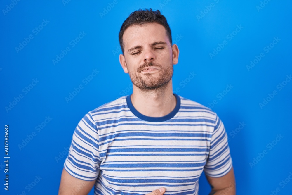 Young hispanic man standing over blue background with hand on stomach because indigestion, painful illness feeling unwell. ache concept. Young hispanic man standing over blue background with hand on stomach because indigestion, painful illness feeling unwell. ache concept.