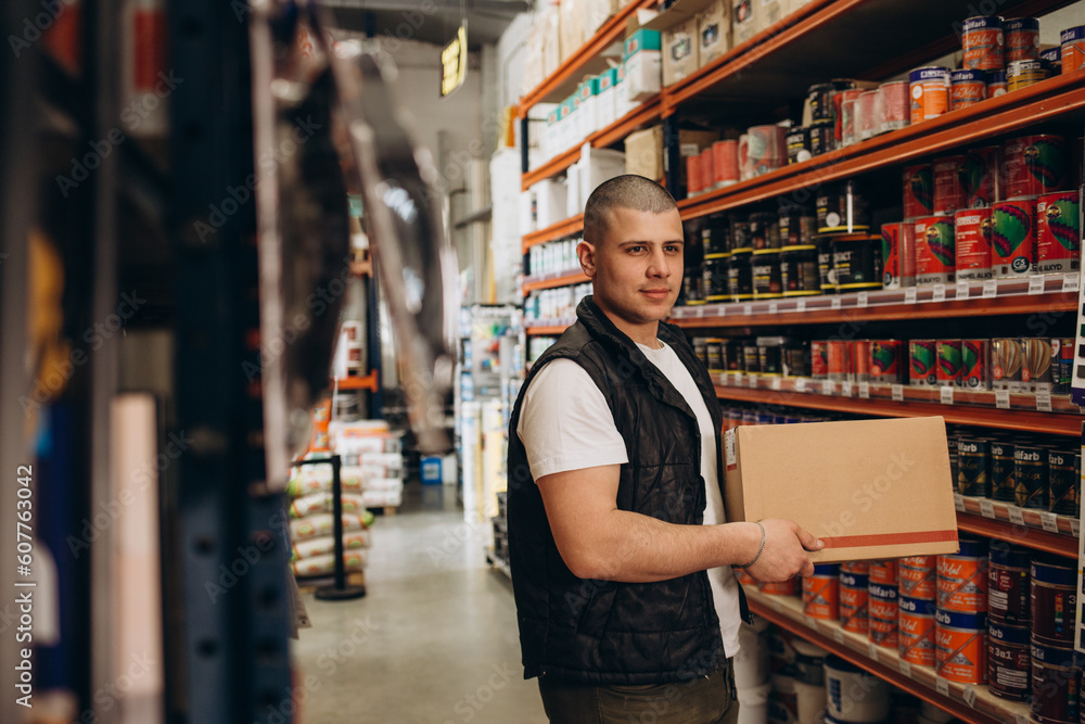 Smiling handsome salesman standing with arms crossed while making eye contact against rack at hardware store. Smiling handsome salesman standing with arms crossed while making eye contact against rack at hardware store.