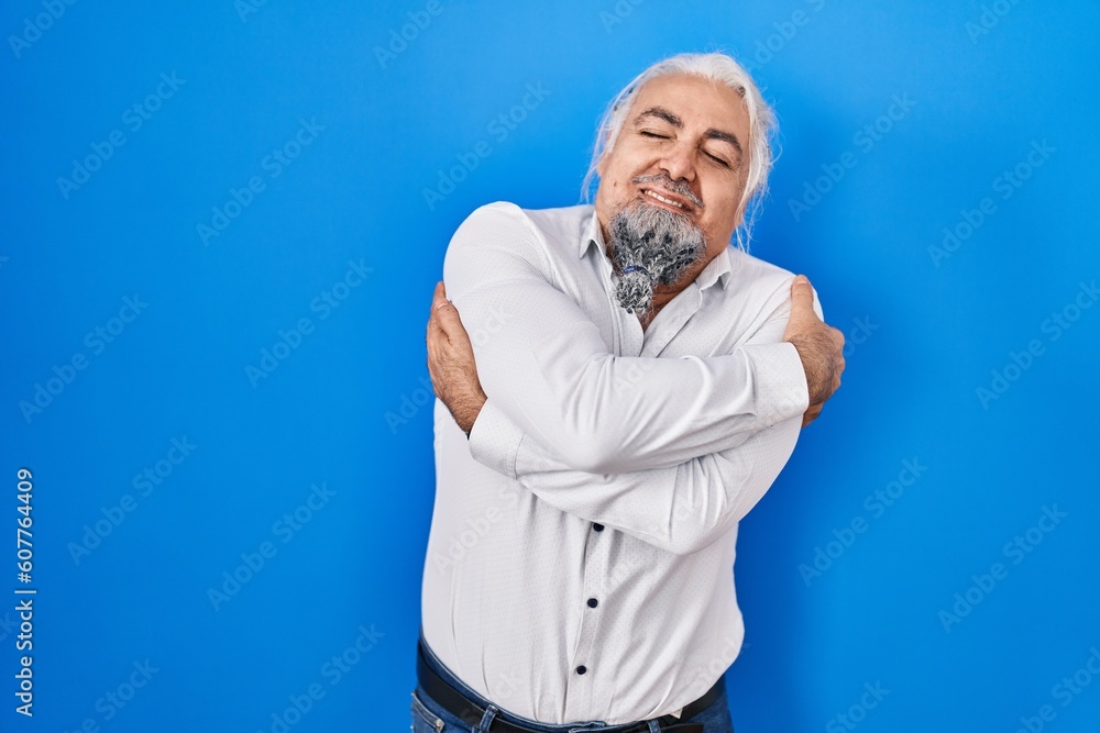 Middle age man with grey hair standing over blue background hugging oneself happy and positive, smiling confident. self love and self care Middle age man with grey hair standing over blue background hugging oneself happy and positive, smiling confident. self love and self care
