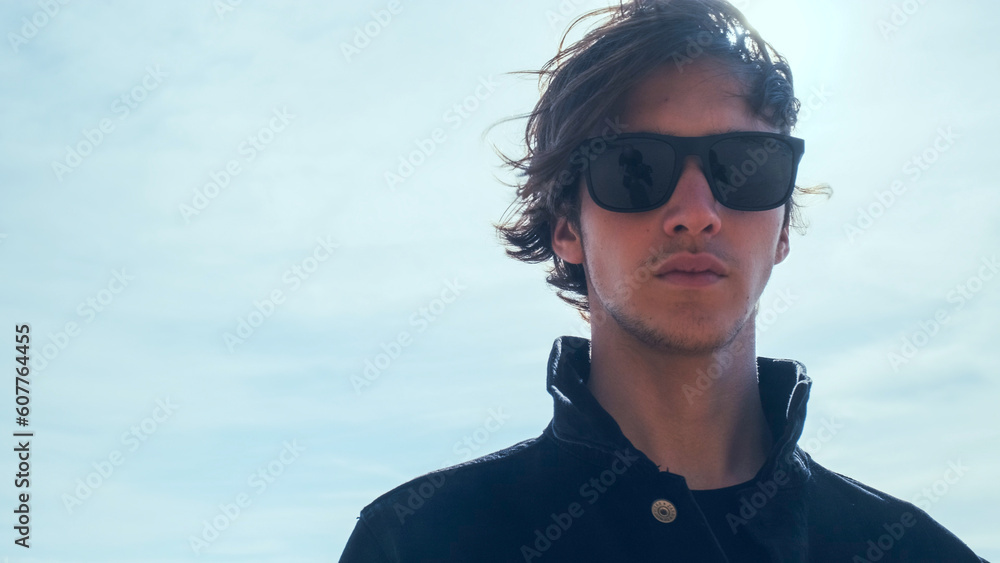 Handsome guy with long hair poses against sky with sun rays. Stylish man in black jacket looks at the camera, low angle shot. Young man with blown brown hair against the blue sky on summer sunny day. Handsome guy with long hair poses against sky with sun rays. Stylish man in black jacket looks at the camera, low angle shot. Young man with blown brown hair against the blue sky on summer sunny day.