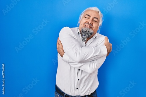 Middle age man with grey hair standing over blue background hugging oneself happy and positive, smiling confident. self love and self care