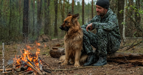 Man with big dog sits at the fire in the forest. Human looks at the fire and touching his dog in nature in autumn. Alone traveler is resting with his German shepherd by the forest fire.