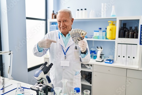 Senior scientist with grey hair working at laboratory holding dollars pointing finger to one self smiling happy and proud