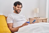Young hispanic man using smartphone and credit card sitting on bed at bedroom