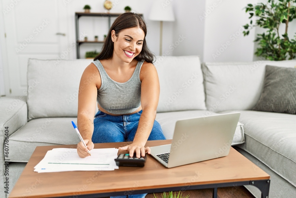 Young beautiful hispanic woman writing on document using calculator at home Young beautiful hispanic woman writing on document using calculator at home