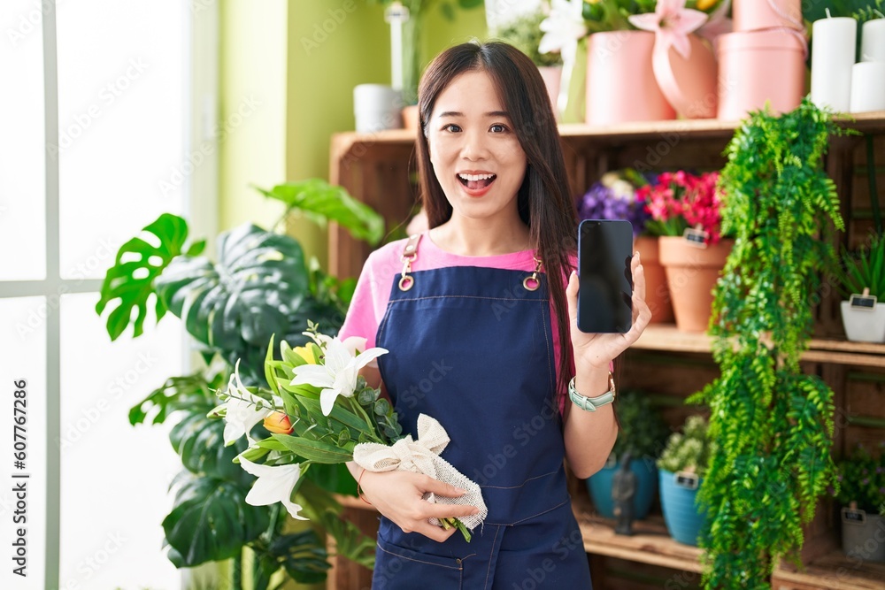 Young chinese woman working at florist shop showing smartphone screen afraid and shocked with surprise and amazed expression, fear and excited face.