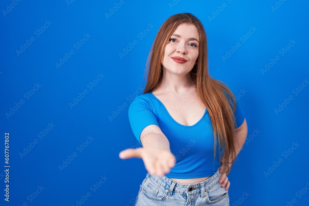 Redhead woman standing over blue background smiling cheerful offering palm hand giving assistance and acceptance. Redhead woman standing over blue background smiling cheerful offering palm hand giving assistance and acceptance.