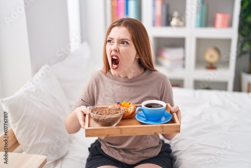 Redhead woman wearing pajama holding breakfast tray angry and mad screaming frustrated and furious, shouting with anger. rage and aggressive concept.