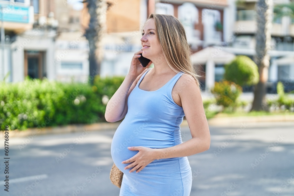 Young pregnant woman talking on smartphone touching belly at street