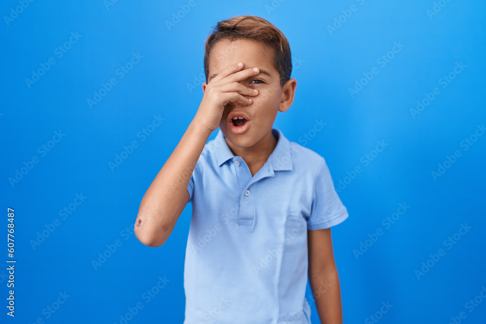 Little hispanic boy wearing casual blue t shirt peeking in shock covering face and eyes with hand, looking through fingers afraid Little hispanic boy wearing casual blue t shirt peeking in shock covering face and eyes with hand, looking through fingers afraid
