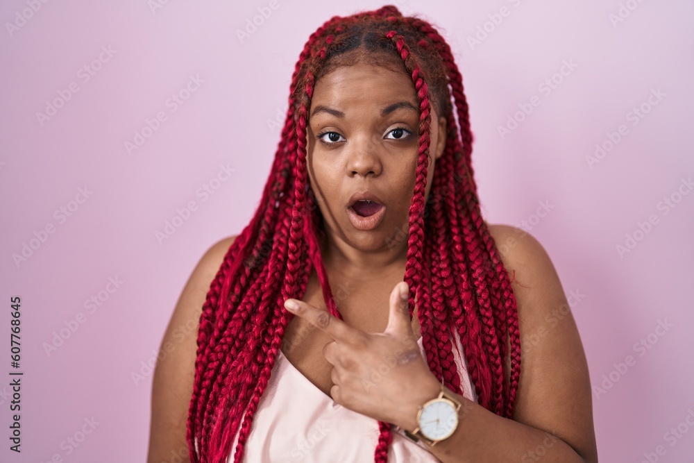African american woman with braided hair standing over pink background surprised pointing with finger to the side, open mouth amazed expression. African american woman with braided hair standing over pink background surprised pointing with finger to the side, open mouth amazed expression.