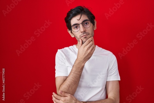 Young hispanic man standing over red background with hand on chin thinking about question, pensive expression. smiling with thoughtful face. doubt concept.