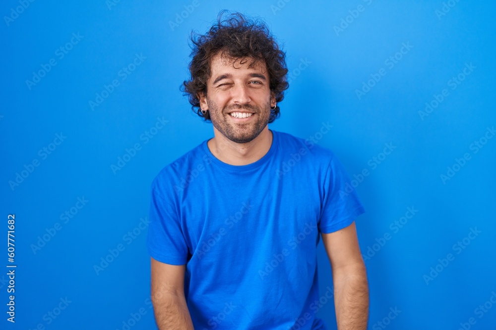 Hispanic young man standing over blue background winking looking at the camera with sexy expression, cheerful and happy face. Hispanic young man standing over blue background winking looking at the camera with sexy expression, cheerful and happy face.