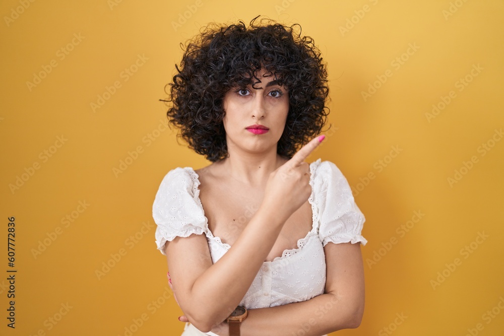 Young brunette woman with curly hair standing over yellow background pointing with hand finger to the side showing advertisement, serious and calm face Young brunette woman with curly hair standing over yellow background pointing with hand finger to the side showing advertisement, serious and calm face