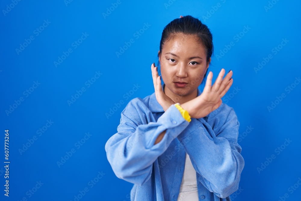 Asian young woman standing over blue background rejection expression crossing arms doing negative sign, angry face Asian young woman standing over blue background rejection expression crossing arms doing negative sign, angry face