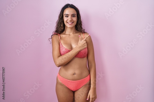 Young hispanic woman wearing lingerie over pink background cheerful with a smile on face pointing with hand and finger up to the side with happy and natural expression