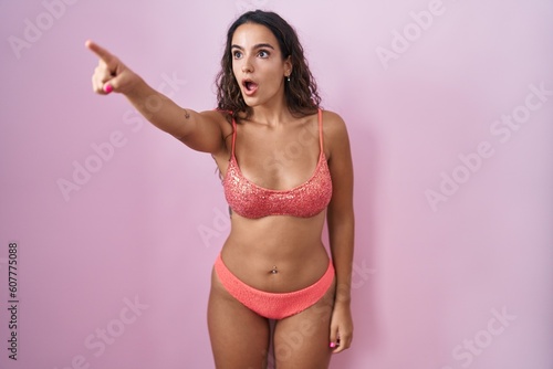 Young hispanic woman wearing lingerie over pink background pointing with finger surprised ahead, open mouth amazed expression, something on the front