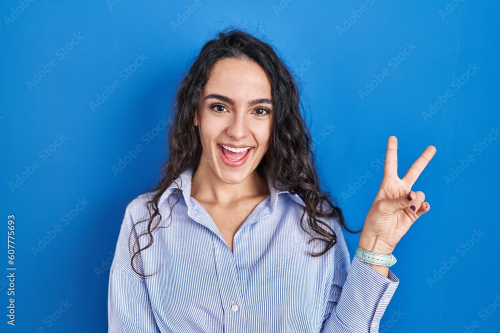 Young brunette woman standing over blue background smiling with happy face winking at the camera doing victory sign. number two. Young brunette woman standing over blue background smiling with happy face winking at the camera doing victory sign. number two.