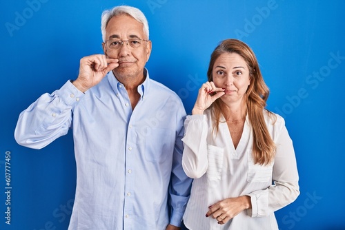Middle age hispanic couple standing over blue background mouth and lips shut as zip with fingers. secret and silent, taboo talking