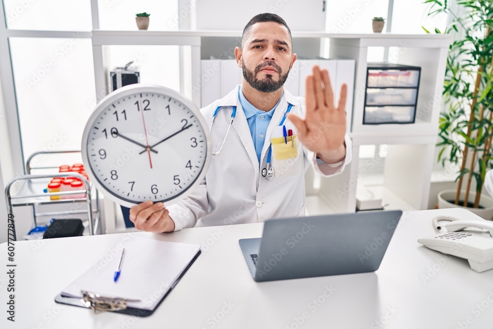 Hispanic doctor man holding clock at the clinic with open hand doing stop sign with serious and confident expression, defense gesture Hispanic doctor man holding clock at the clinic with open hand doing stop sign with serious and confident expression, defense gesture