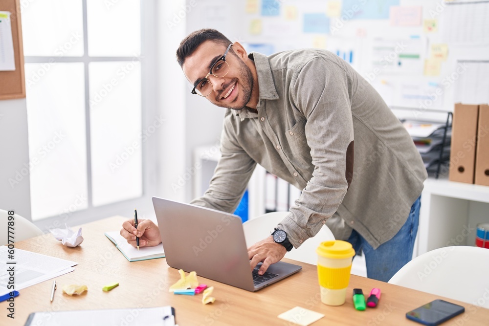 Young hispanic man business worker using laptop writing on notebook at office Young hispanic man business worker using laptop writing on notebook at office