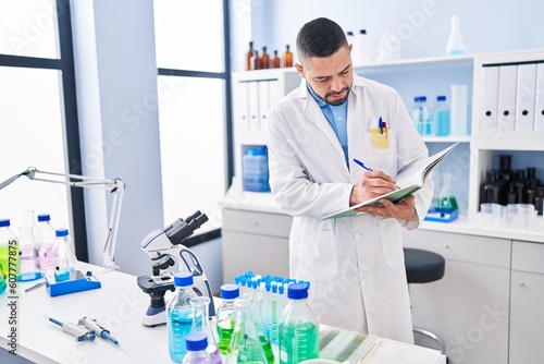 Young latin man scientist writing on notebook at laboratory