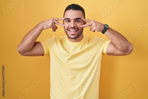 Young hispanic man standing over yellow background doing peace symbol with fingers over face, smiling cheerful showing victory