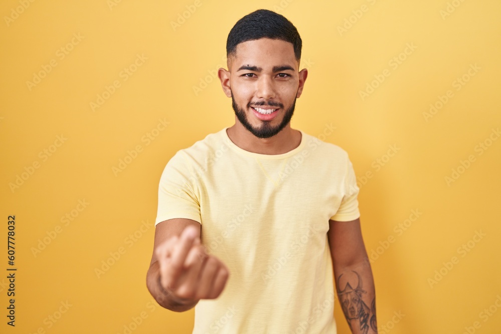 Young hispanic man standing over yellow background beckoning come here gesture with hand inviting welcoming happy and smiling Young hispanic man standing over yellow background beckoning come here gesture with hand inviting welcoming happy and smiling