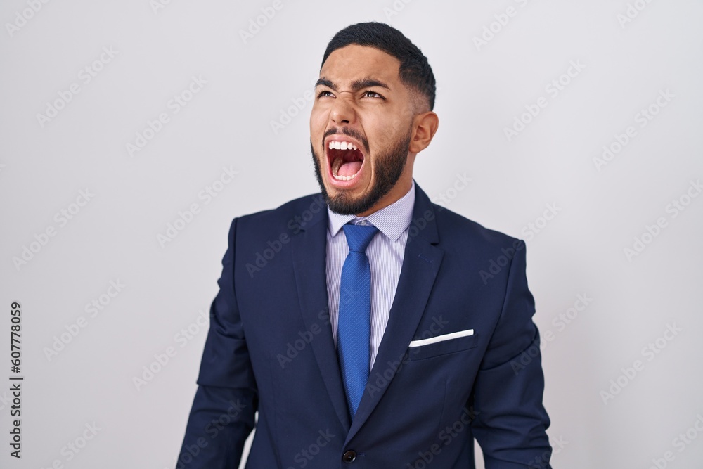 Young hispanic man wearing business suit and tie angry and mad screaming frustrated and furious, shouting with anger. rage and aggressive concept. Young hispanic man wearing business suit and tie angry and mad screaming frustrated and furious, shouting with anger. rage and aggressive concept.