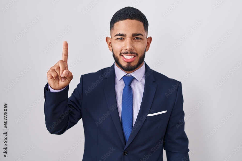 Young hispanic man wearing business suit and tie showing and pointing up with finger number one while smiling confident and happy. Young hispanic man wearing business suit and tie showing and pointing up with finger number one while smiling confident and happy.
