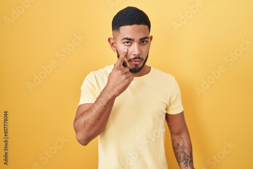Young hispanic man standing over yellow background pointing to the eye watching you gesture, suspicious expression © Krakenimages.com