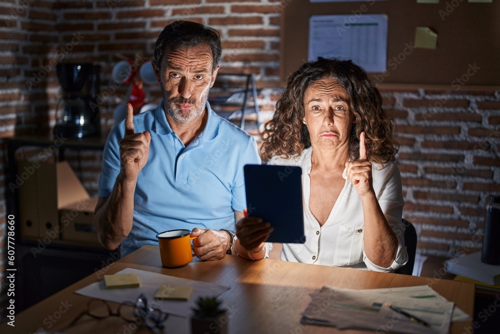 Middle age hispanic couple using touchpad sitting on the table at night pointing up looking sad and upset, indicating direction with fingers, unhappy and depressed. Middle age hispanic couple using touchpad sitting on the table at night pointing up looking sad and upset, indicating direction with fingers, unhappy and depressed.