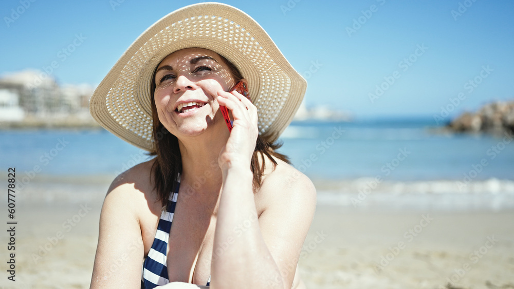 Middle age hispanic woman tourist sunbathing sitting on the towel speaking on the phone at the beach