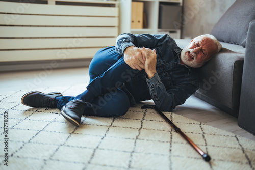 Elder senior man lying on floor after falling down with wooden walking stick beside couch on rug in living room at home. Old man suffering with pain and struggling to get up after falling down at home © Dragana Gordic