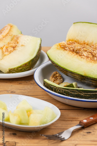 View of half a green melon, plate with pieces and fork on wooden table, white background, vertical, with copy space